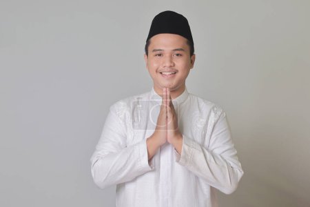 Portrait of Asian muslim man in white koko shirt showing apologize and welcome hand gesture. Isolated image on gray background