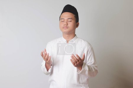 Porträt eines asiatischen muslimischen Mannes in weißem Kokohemd mit Totenkopf, der mit erhobenen Händen ernsthaft betet und aufblickt. Isoliertes Bild auf grauem Hintergrund