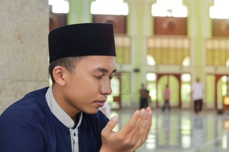Portrait de religieux asiatique en chemise musulmane et casquette noire priant avec la main levée à l'intérieur de la mosquée publique