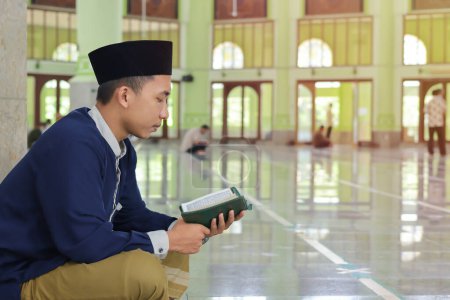 Photo for Religious Asian man in muslim shirt and black cap reading the holy book of Quran in the public mosque - Royalty Free Image