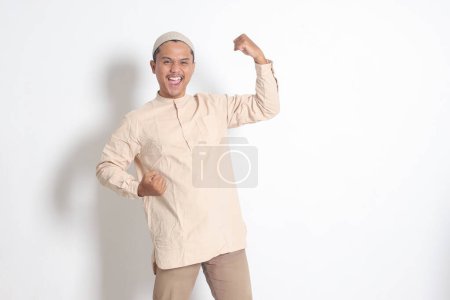 Portrait of religious Asian muslim man in koko shirt with skullcap raising his fist, celebrating success. Achievement concept. Isolated image on white background