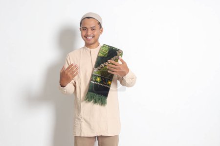Téléchargez les photos : Portrait d'homme musulman asiatique séduisant en chemise koko avec bonnet de crâne invitant et accueillant quelqu'un à entrer. Image isolée sur fond blanc - en image libre de droit