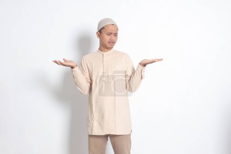 Porträt eines verwirrten asiatischen muslimischen Mannes im Kokohemd mit Totenkopf, der die Hände seitlich spreizt und zwei Dinge hält, um Produkte zu demonstrieren. Isoliertes Bild auf weißem Hintergrund