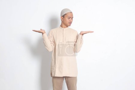 Porträt eines verwirrten asiatischen muslimischen Mannes im Kokohemd mit Totenkopf, der die Hände seitlich spreizt und zwei Dinge hält, um Produkte zu demonstrieren. Isoliertes Bild auf weißem Hintergrund
