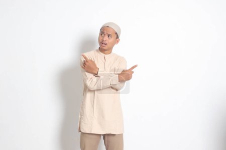 Porträt eines aufgeregten asiatischen muslimischen Mannes im Kokohemd mit zur Seite zeigender Schirmmütze, der die Wahl zwischen zwei Objekten trifft, das Produkt demonstriert, die Hände verschränkt. Isoliertes Bild auf weißem Hintergrund