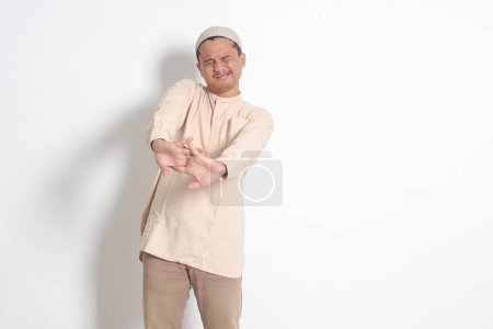 Portrait of overworked Asian muslim man in koko shirt with skullcap stretching his hands and body after waking up. Sleep deprivation concept. Isolated image on white background