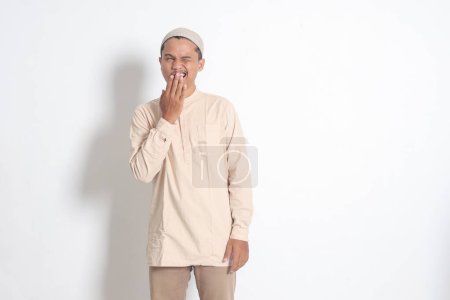 Portrait of overworked Asian muslim man in koko shirt with skullcap yawning with hand covering his mouth. Sleep deprivation concept. Isolated image on white background