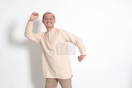 Portrait of overworked Asian muslim man in koko shirt with skullcap stretching his hands and body after waking up. Sleep deprivation concept. Isolated image on white background