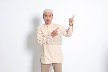 Portrait of shocked Asian muslim man in koko shirt with skullcap showing product and pointing with his hand and finger to the side. Advertising concept. Isolated image on white background