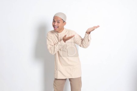 Porträt eines schockierten asiatischen muslimischen Mannes im Kokohemd mit Totenkopf, der das Produkt zeigt und mit der Hand und dem Finger zur Seite zeigt. Werbekonzept. Isoliertes Bild auf weißem Hintergrund