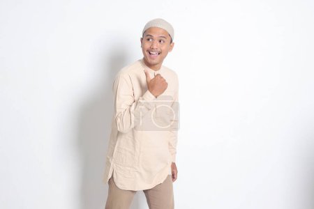 Portrait of shocked Asian muslim man in koko shirt with skullcap showing product and pointing with thumb up to the side. Advertising concept. Isolated image on white background
