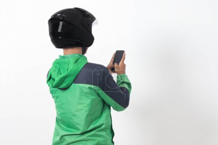 Téléchargez les photos : Vue arrière du chauffeur de taxi en ligne asiatique portant une veste verte et un casque tenant un téléphone portable avec écran blanc. Image isolée sur fond blanc - en image libre de droit