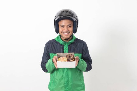 Téléchargez les photos : Portrait de chauffeur de taxi en ligne asiatique portant une veste verte et un casque tenant une boîte de beignets, livrant au client. Image isolée sur fond blanc - en image libre de droit