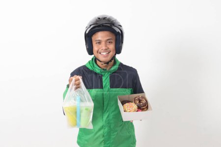 Téléchargez les photos : Portrait d'un chauffeur de taxi en ligne asiatique portant une veste verte et un casque tenant une boîte de beignets et une tasse de boisson jus, livrant au client. Image isolée sur fond blanc - en image libre de droit