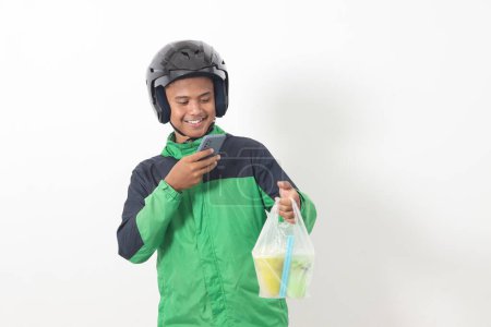 Portrait of Asian online taxi driver wearing green jacket and helmet delivering the beverages in plastic cup to customer, while using mobile phone. Isolated image on white background