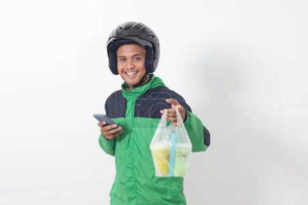 Téléchargez les photos : Portrait d'un chauffeur de taxi asiatique en ligne portant une veste verte et un casque livrant les boissons dans une tasse en plastique au client, tout en utilisant un téléphone mobile. Image isolée sur fond blanc - en image libre de droit