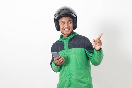 Téléchargez les photos : Portrait d'un chauffeur de taxi asiatique en ligne portant une veste verte et un casque tenant un téléphone portable tout en pointant vers le côté. Image isolée sur fond blanc - en image libre de droit
