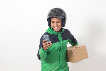 Téléchargez les photos : Portrait de chauffeur de messagerie en ligne asiatique portant une veste verte et un casque livrant le paquet et la boîte pour le client tout en utilisant le téléphone mobile et en prenant commande. Image isolée sur fond blanc - en image libre de droit