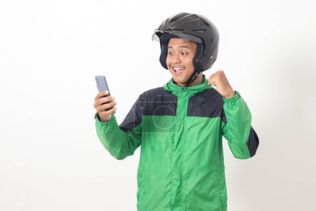 Téléchargez les photos : Portrait de chauffeur de taxi en ligne asiatique portant une veste verte et un casque à l'aide d'un téléphone portable tout en levant le poing et en célébrant le succès. Image isolée sur fond blanc - en image libre de droit