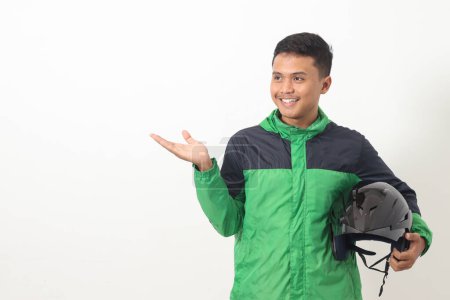 Téléchargez les photos : Portrait de chauffeur de taxi en ligne asiatique portant vert debout sur fond blanc, tenant son casque sur sa hanche tout en pointant vers le côté - en image libre de droit