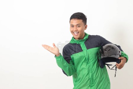 Téléchargez les photos : Portrait de chauffeur de taxi en ligne asiatique portant vert debout sur fond blanc, tenant son casque sur sa hanche tout en pointant vers le côté - en image libre de droit