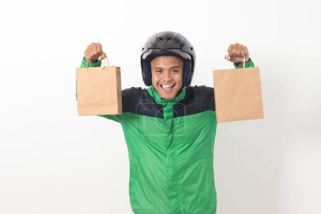 Téléchargez les photos : Portrait de chauffeur de messagerie en ligne asiatique portant une veste verte et un casque donnant sac à provisions en papier artisanal avec cadeau à l'intérieur. Image isolée sur fond gris - en image libre de droit