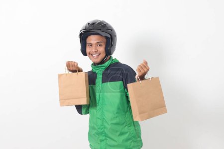 Téléchargez les photos : Portrait de chauffeur de messagerie en ligne asiatique portant une veste verte et un casque donnant sac à provisions en papier artisanal avec cadeau à l'intérieur. Image isolée sur fond gris - en image libre de droit