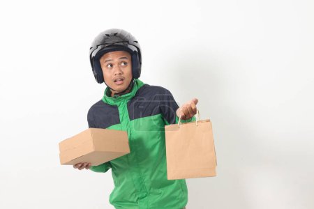 Téléchargez les photos : Portrait de chauffeur de messagerie en ligne asiatique portant une veste verte et un casque donnant sac à provisions en papier artisanal et paquet boîte. Image isolée sur fond blanc - en image libre de droit
