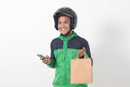 Téléchargez les photos : Portrait de chauffeur de messagerie en ligne asiatique portant une veste verte et un casque donnant sac à provisions en papier artisanal tout en utilisant un téléphone mobile. Image isolée sur fond blanc - en image libre de droit