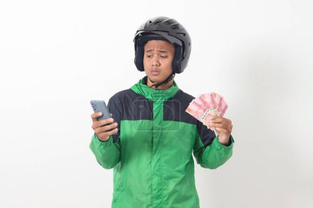 Portrait of Asian online taxi driver wearing green standing against white background, smiling and looking at camera, holding a bunch of rupiah money