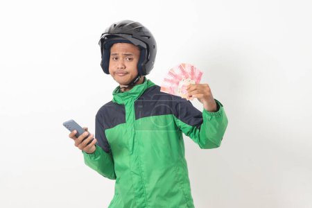 Téléchargez les photos : Portrait de chauffeur de taxi en ligne asiatique portant vert debout sur fond blanc, souriant et regardant la caméra, tenant un tas d'argent rupiah - en image libre de droit