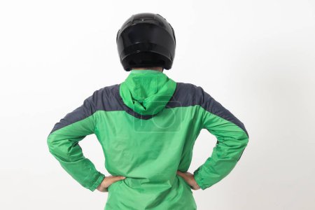 Téléchargez les photos : Vue arrière du chauffeur de messagerie en ligne asiatique portant une veste verte et un casque plaçant les mains sur la hanche, se sentant fatigué. Image isolée sur fond blanc - en image libre de droit