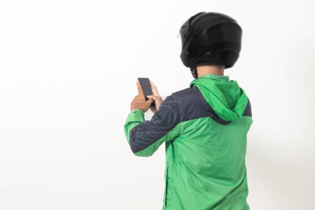 Téléchargez les photos : Vue arrière du chauffeur de taxi en ligne asiatique portant une veste verte et un casque tenant un téléphone portable avec écran blanc. Image isolée sur fond blanc - en image libre de droit
