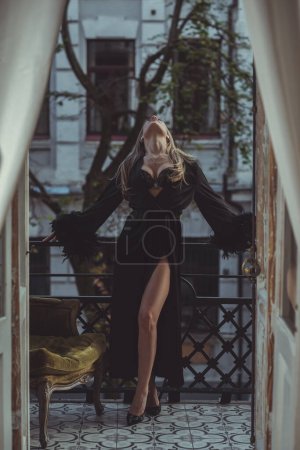 Foto de Dama en peignoir con una copa de vino. Foto sensual de mujer rubia europea en bata de seda negra con mangas de plumas. Concepto de moda - Imagen libre de derechos
