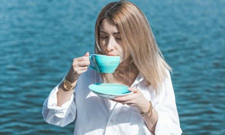 Modern European woman of middle age in white shirt with a blue cup of coffee at morning sunny day relaxing