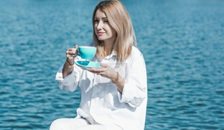 Modern European woman of middle age in white shirt with a blue cup of coffee at morning sunny day relaxing