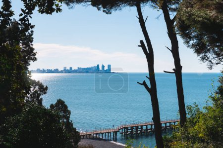 View of downtown Batumi from the nearby beach of the Botanical Garden with a pier, framed by trees and greenery against a clear blue sky and calm sea