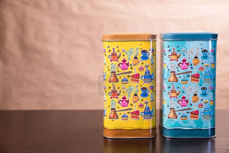 Two multi-colored metal cans for bulk products, cookies, sugar or tea with a pattern on the theme of tea drinking. Yellow and turquoise on a beige background