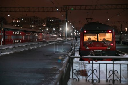 Estación de tren de invierno nocturno. Trenes rojos en la plataforma. Silueta de una persona, faroles y edificios de gran altura en la distancia