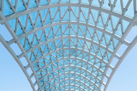 Una estructura suspendida hecha de tubos de metal blanco y vidrio en forma de arcos e intersecciones contra un cielo despejado