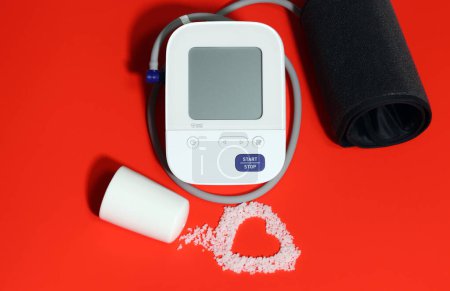 Electronic blood pressure gauge and electrocardiogram-shaped salt on a red background. Eating salt can increase high blood pressure and heart disease.