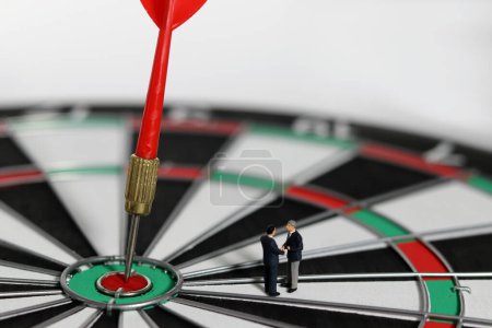 Business goals and concepts for achievement and victory. Miniature entrepreneurs shaking hands over a dart board with a red dart arrow stuck in the center.