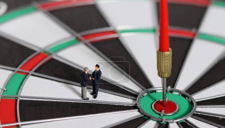 Business goals and concepts for achievement and victory. Miniature entrepreneurs shaking hands over a dart board with a red dart arrow stuck in the center.