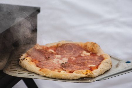 Téléchargez les photos : Cuisson ou cuisson de pizza chaude avec sauce tomate, mozzarella et salami avec une pelle dans un four électrique ou à gaz, avec de la fumée, gros plan - en image libre de droit