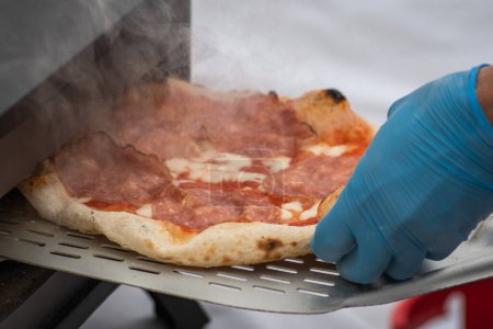 Téléchargez les photos : Cuisson ou cuisson avec un gant en plastique pizza chaude avec sauce tomate, mozzarella et salami viande avec une pelle dans un four électrique ou à gaz, avec de la fumée, fermer - en image libre de droit