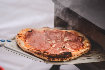 Téléchargez les photos : Cuisson ou cuisson de pizza chaude avec sauce tomate, mozzarella et salami avec une pelle dans un four électrique ou à gaz, avec de la fumée, gros plan - en image libre de droit