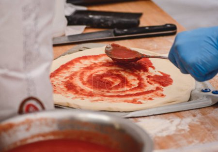 Téléchargez les photos : Ajouter la sauce tomate avec une cuillère sur la pâte à pizza sur une pelle sur une planche en bois, fermer - en image libre de droit