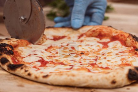 Photo for Hand with plastic glove cutting hot pizza Margherita with tomato sauce and mozzarella cheese with a cutter on a wooden board, close up - Royalty Free Image