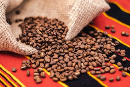Téléchargez les photos : Roasted coffee beans or seeds in a jute sack and scattered on a tablecloth with African decorations, close up - en image libre de droit