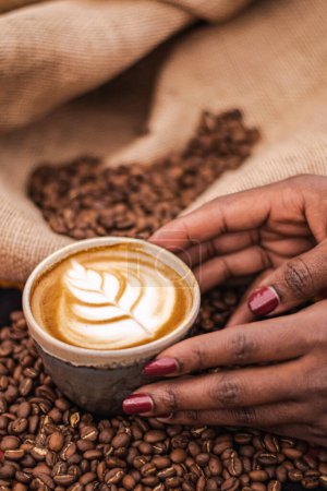 Téléchargez les photos : Black skin girl holding a Cappuccino in a ceramic cup with flower or leaves drawn on the foam, teapot and roasted coffee beans or seeds in a jute sack, vertical - en image libre de droit